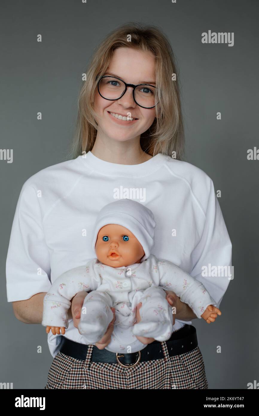 Portrait of young happy beauteous woman holding baby doll in white clothes with two hands on grey background. Studio. Stock Photo