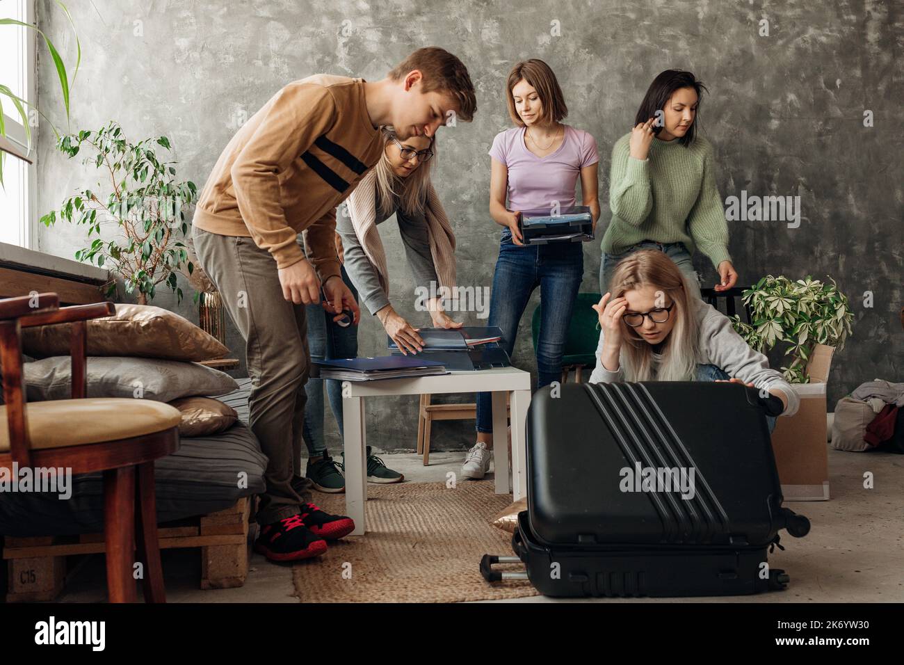 Smiling, busy friends pack luggage, box with pillows, plants, clothes in living room, show out female friend to moving Stock Photo