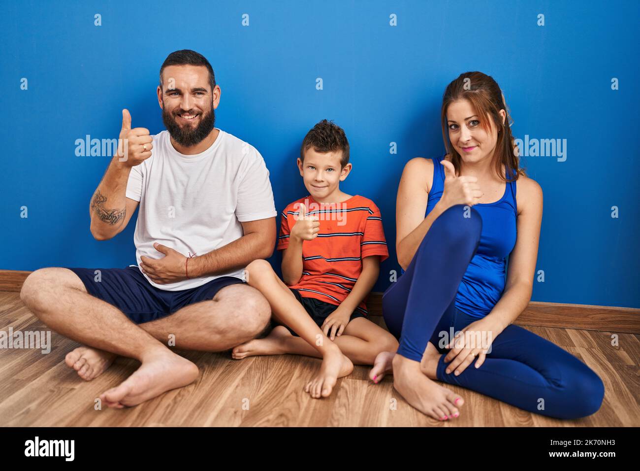 Family of three sitting on the floor at home doing happy thumbs up gesture with hand. approving expression looking at the camera showing success. Stock Photo