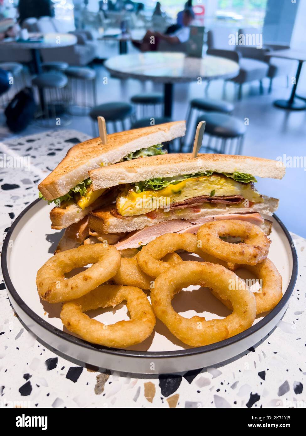 Vertical view of a large size club sandwich with marinated chicken, turkey bacon, ham, scrambled eggs and onion rings as the side. Stock Photo