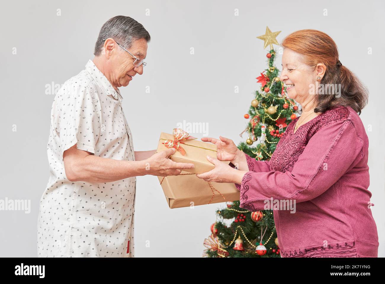 Senior latin couple smiling, spending the holidays at home, exchanging Christmas gifts near a decorated tree. The joy of celebrating together. Stock Photo