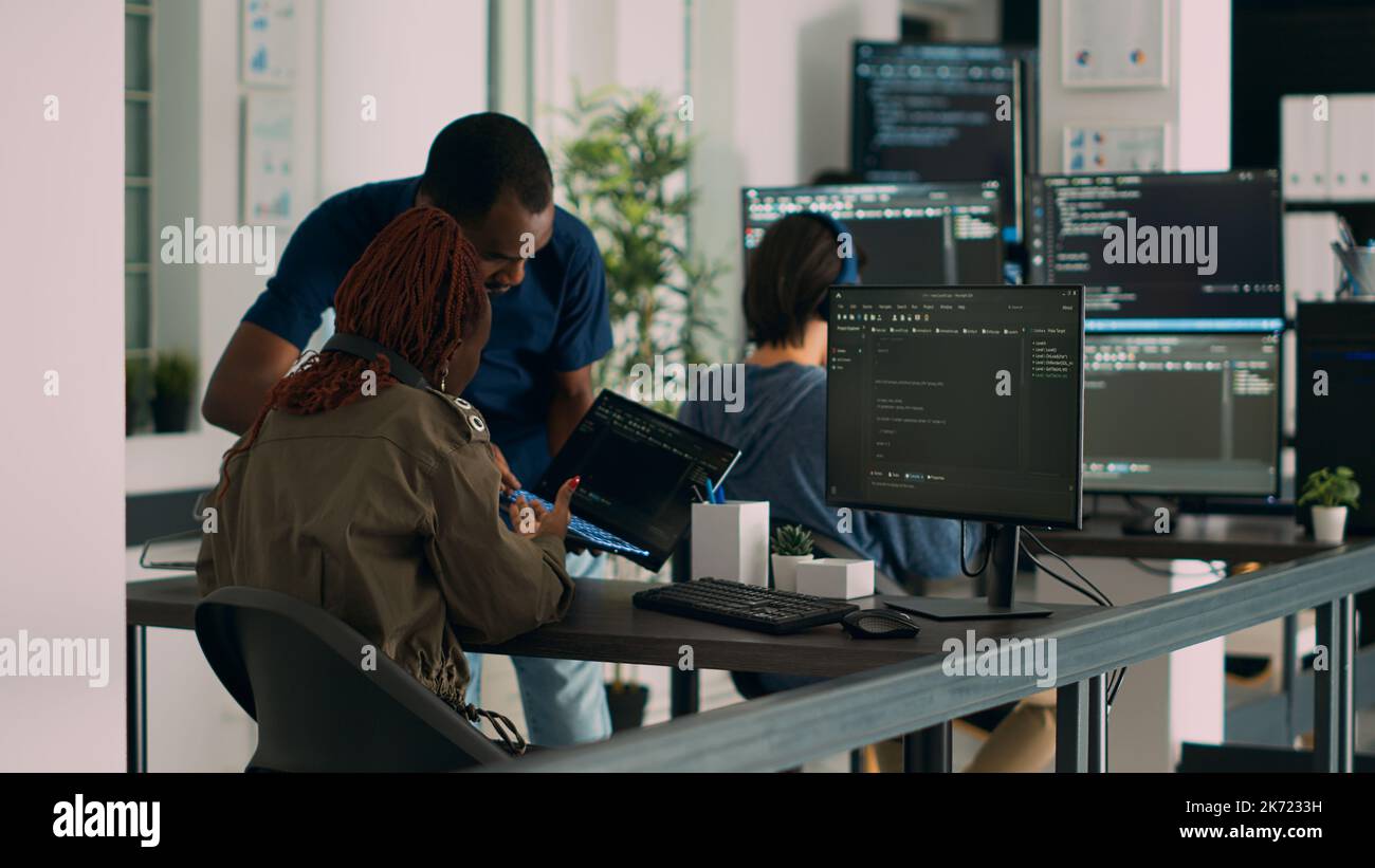 Database developer explaining algorithm to colleague coder pointing at running code on laptop. System engineers collaborating on development group project, using terminal window. Stock Photo