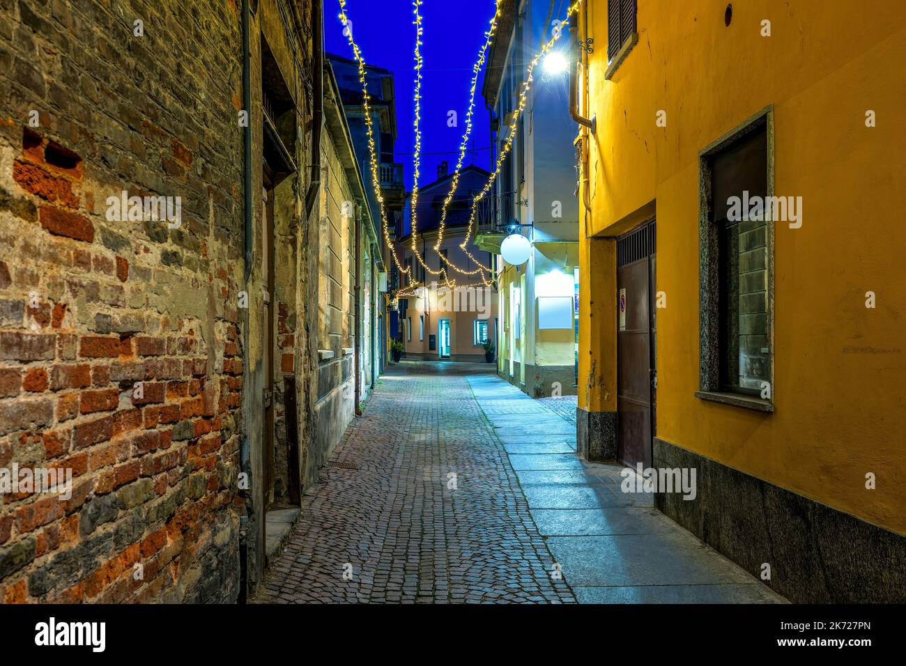 Narrow cobblestone street among old houses illuminated with Christmas lights in evening in Alba, Piedmont, Northern Italy. Stock Photo
