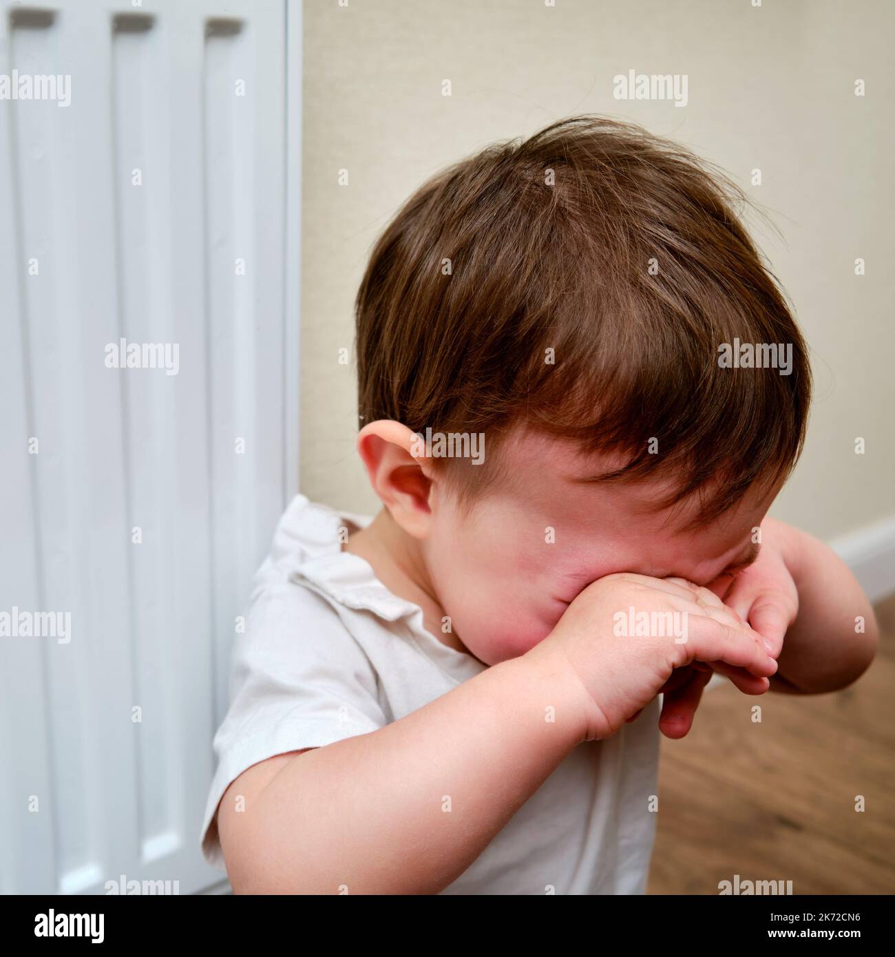 Toddler baby crying at the radiator, tearful child face closeup. White