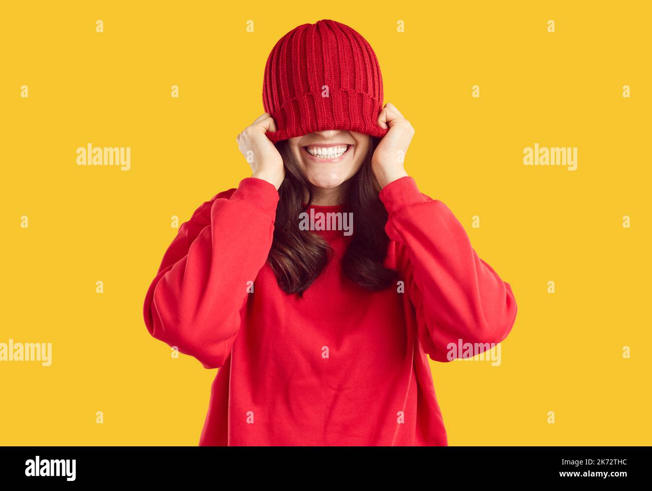 Joyful young woman covers her eyes with warm red winter hat that she puts on her head Stock Photo