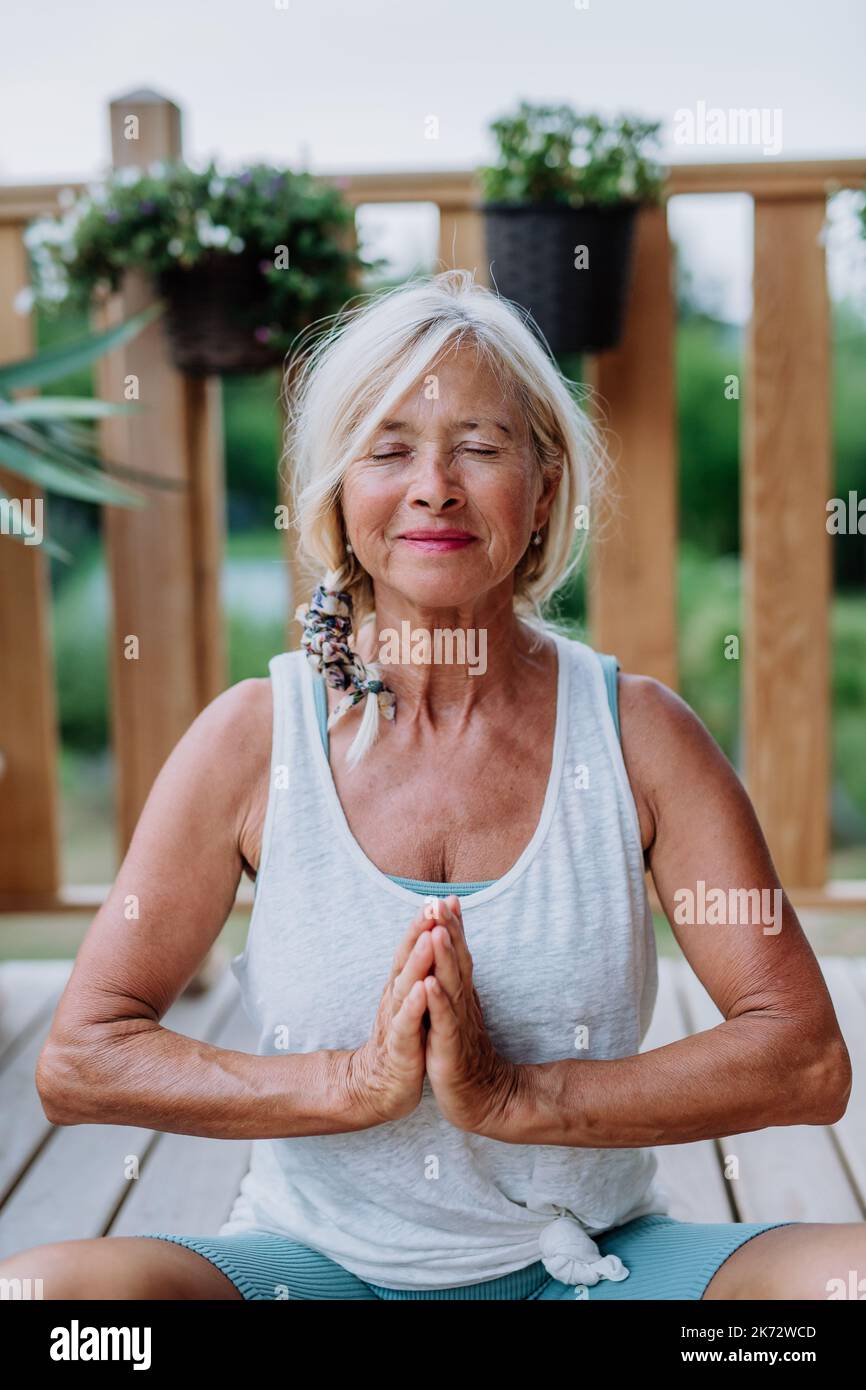 A senior woman sitting outdoors on a terrace in summer, doing yoga ...