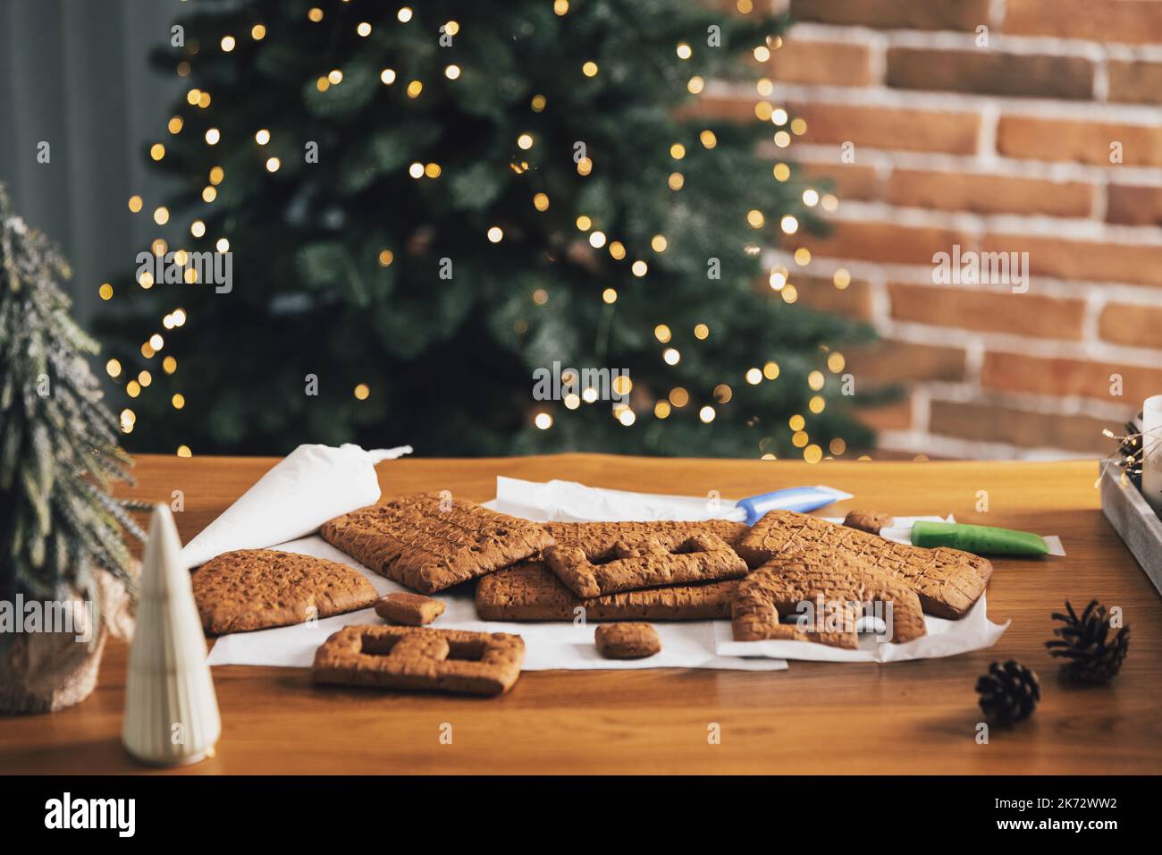 Homemade gingerbread house building blocks with glaze laying on table with decorations, lanterns in living room decorated with defocused New Year lights, Christmas fir tree. Holiday mood for kids Stock Photo