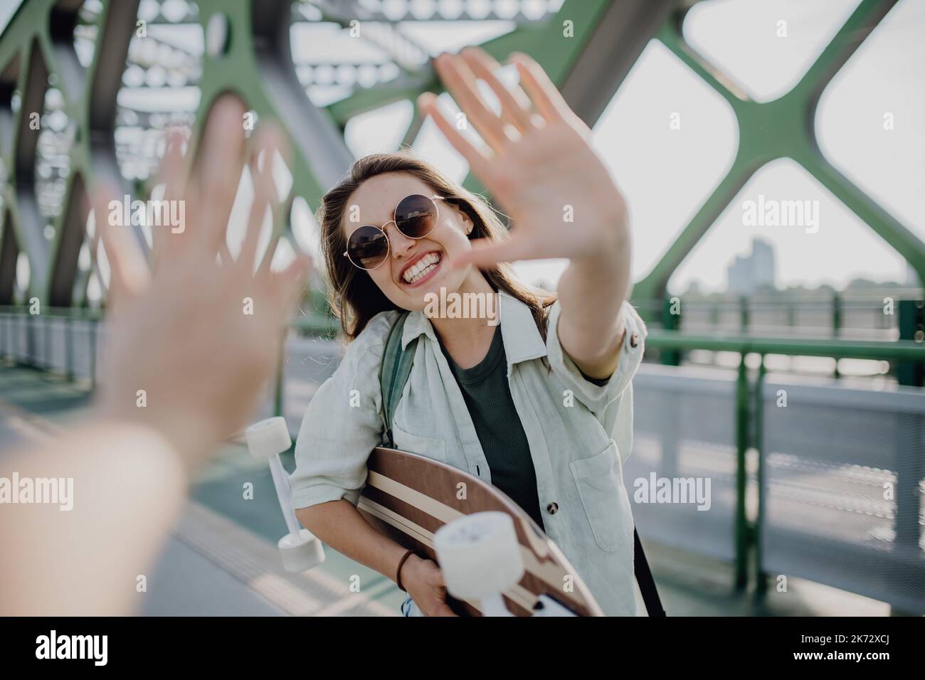 Young woman on city bridge with skateboard greating someone. Youth culture and commuting concept. Stock Photo
