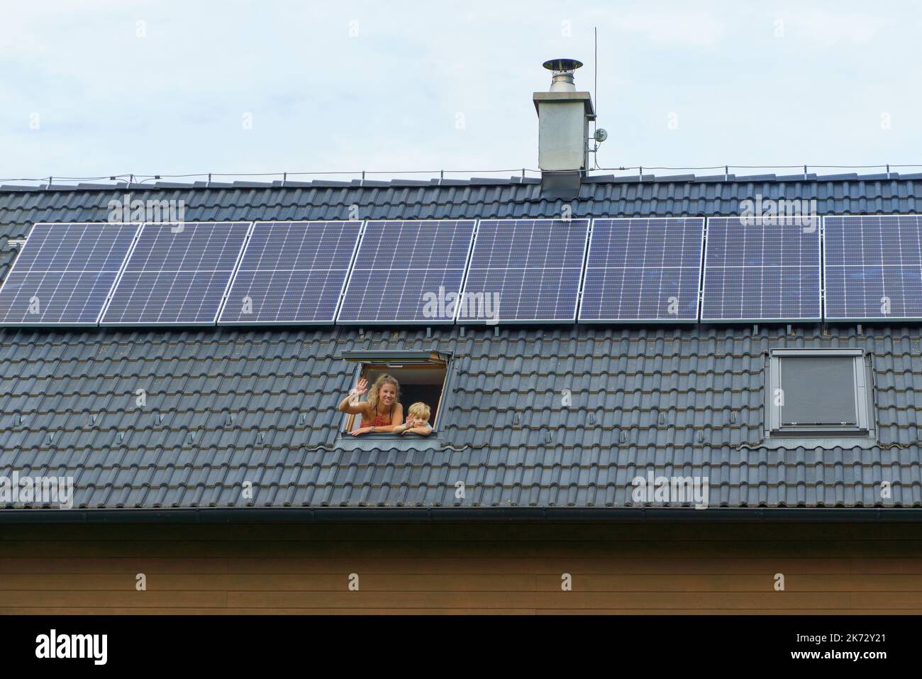 Happy mother and her son waving from skylight window in their new house with solar panels on the roof. Alternative energy, saving resources and Stock Photo