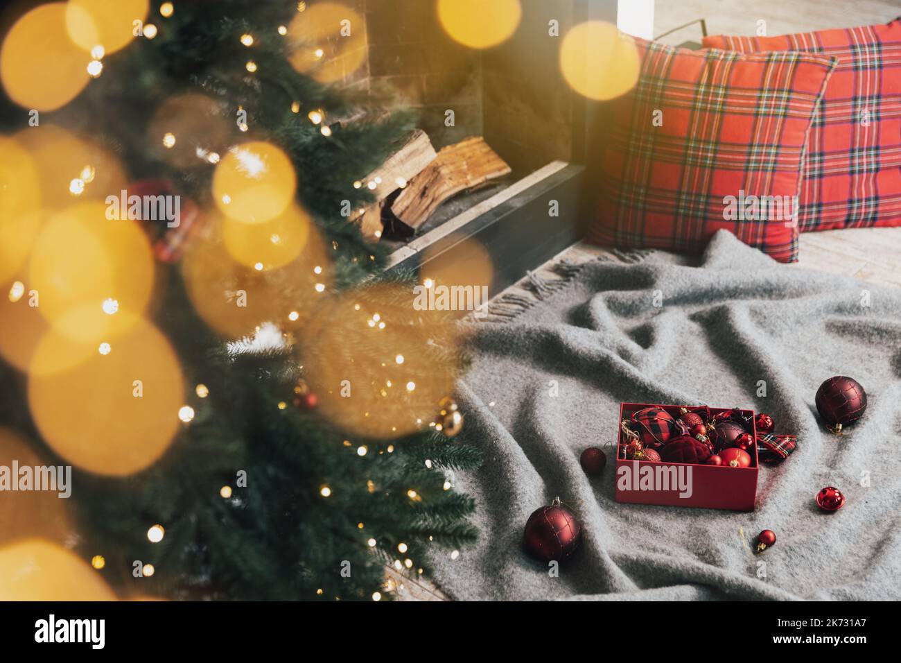 Box of Christmas balls laying on gray carpet or plaid ready for use for decorating and hanging on green Christmas fir tree with lights, garland in the living room near fireplace with wood inside Stock Photo