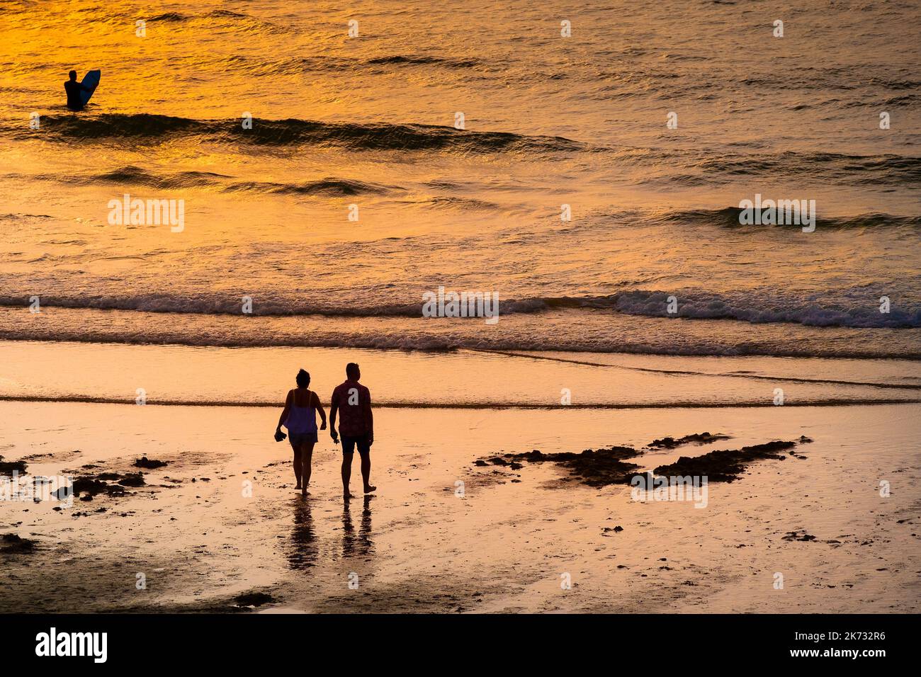 Two holidaymakers silhouetted walking to the shoreline on Fistral Beach at sunset in Newquay in Cornwall in the UK in Europe. Stock Photo