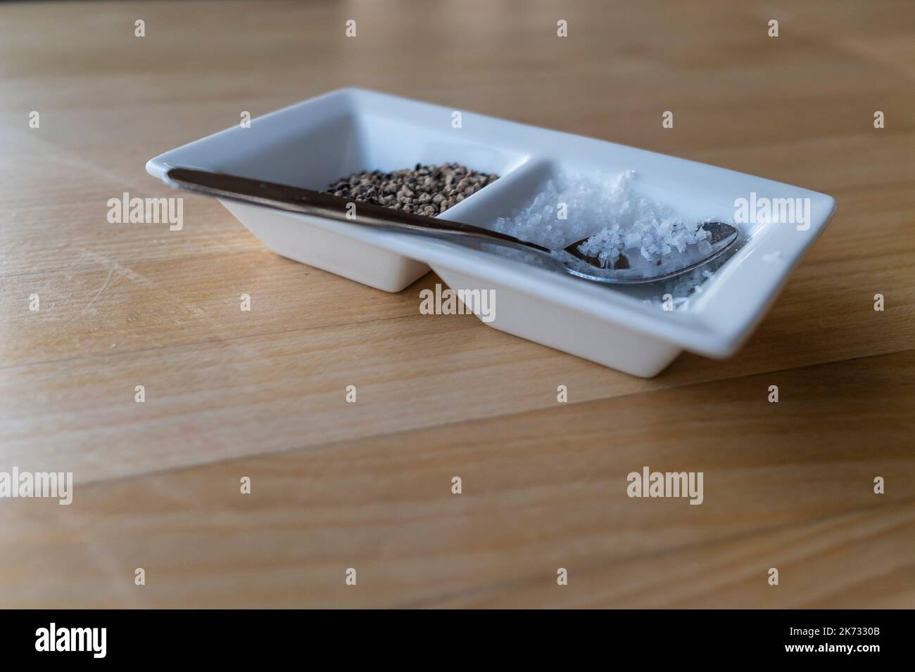 A close up closeup view of a spoon resting on a small dish of sea salt and cracked black pepper on a table. Stock Photo