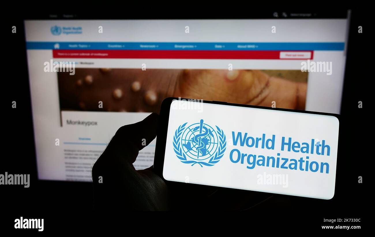 Person holding mobile phone with logo of UN agency World Health Organization (WHO) on screen in front of web page. Focus on phone display. Stock Photo