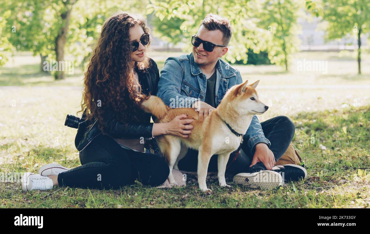 Husband and wife happy couple are fussing their pet shiba inu puppy sitting on grass in park and talking. Conversation, domestic animals and modern lifestyle concept. Stock Photo