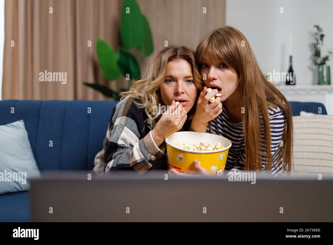 Two women of different ages watch horror movie and eat popcorn while sitting on the couch hugging each other Stock Photo