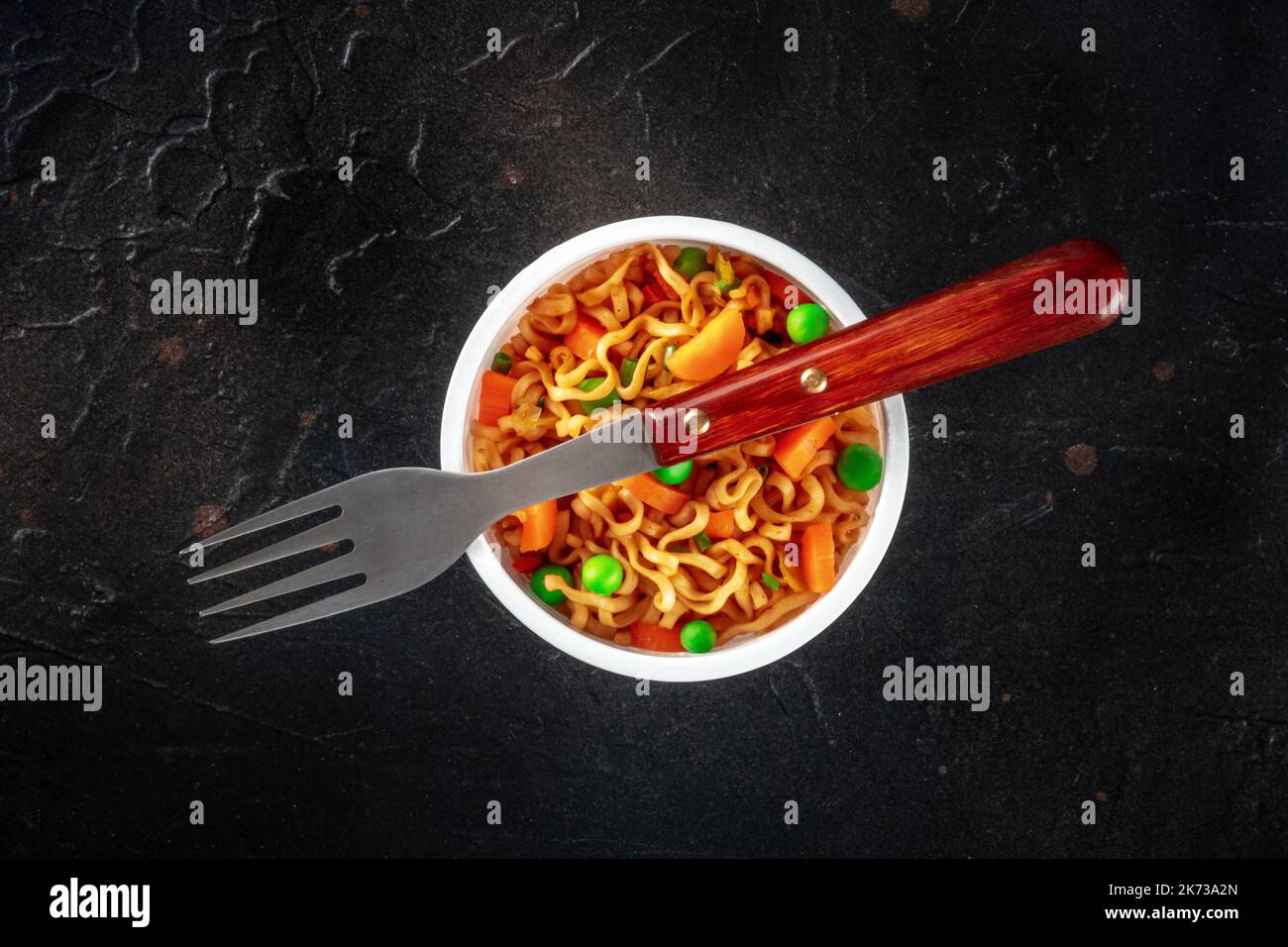Ramen cup, instant soba noodles in a plastic cup with vegetables, green peas, onions, and carrots, top shot with a fork on a black slate background Stock Photo