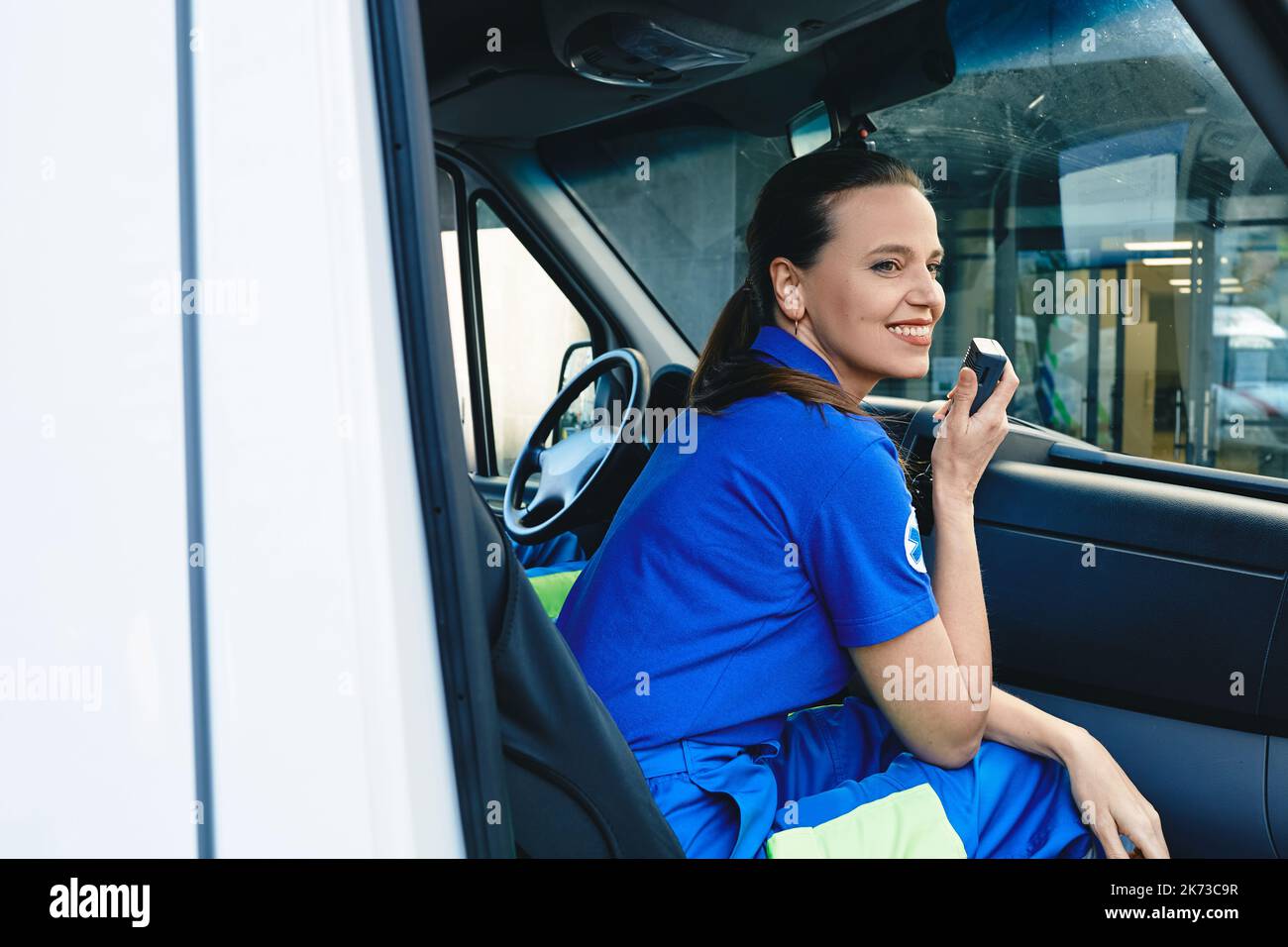 Emergency medical services worker. Smiling female paramedic talking on portable radio while sitting in ambulance Stock Photo