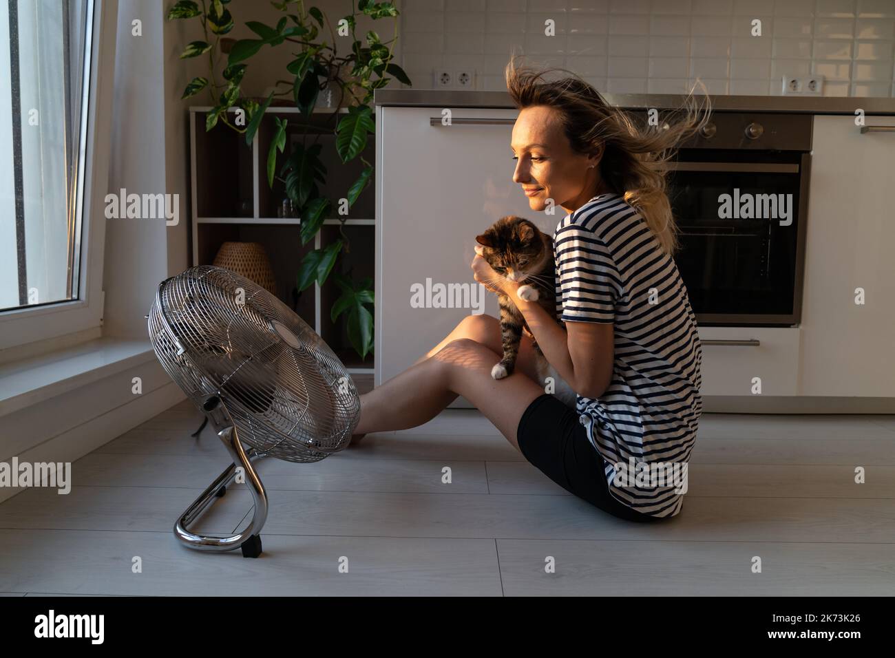 Young woman sitting near electric fan helps cat to cool down from
