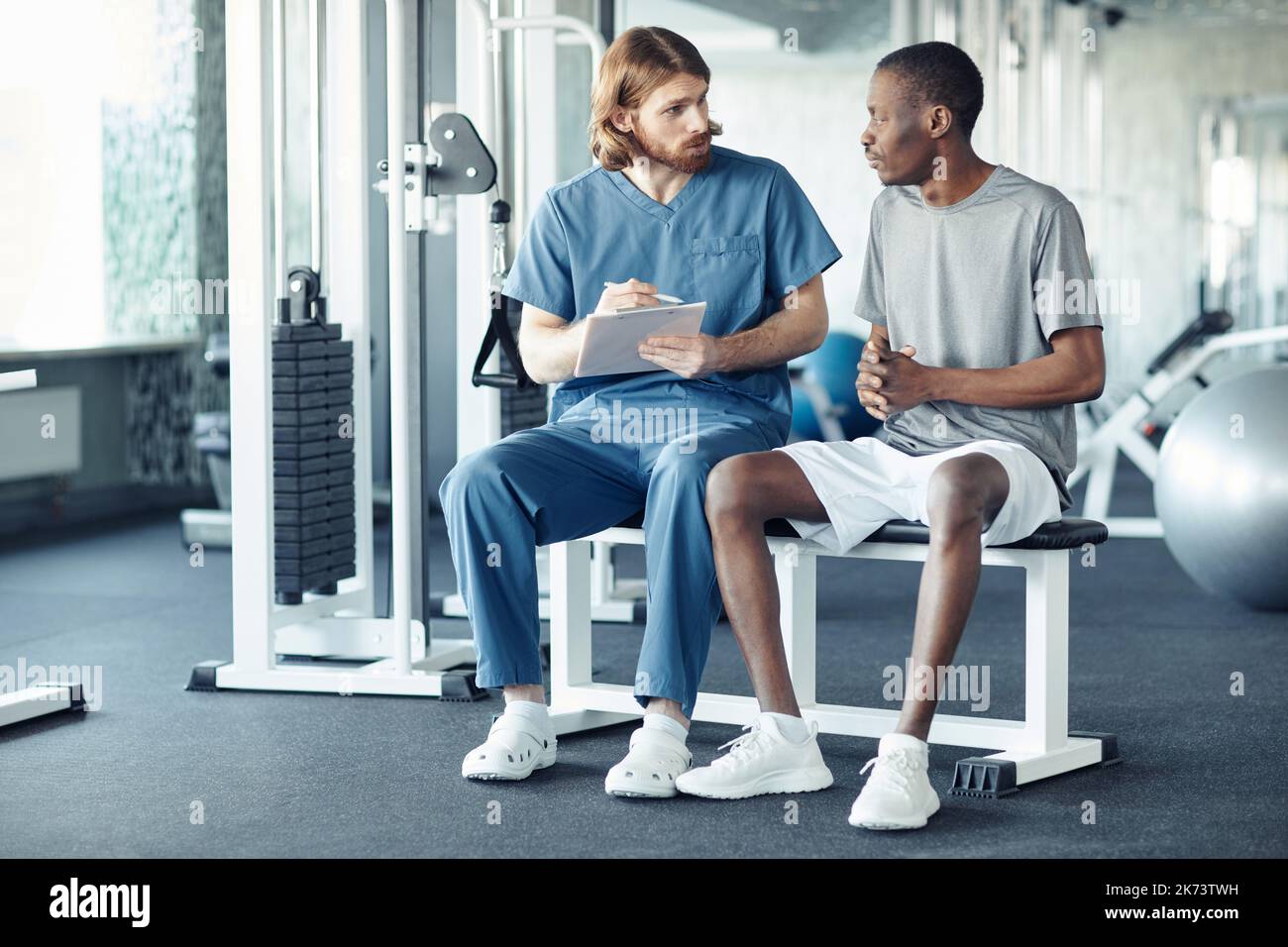 Young doctor asking questions to patient about his rehabilitation while they sitting in gym Stock Photo