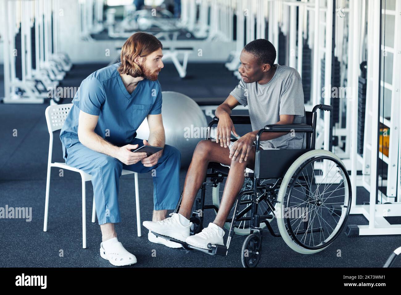 Young doctor in uniform discussing rehabilitation with patient while he sitting in wheelchair in gym Stock Photo