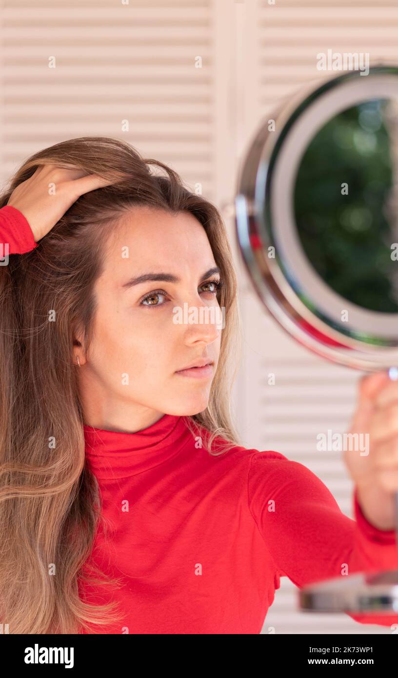 Vertical view of young woman suffering from alopecia. Female hair loss.Worried female looking her hairline in the mirror in red outfit Stock Photo