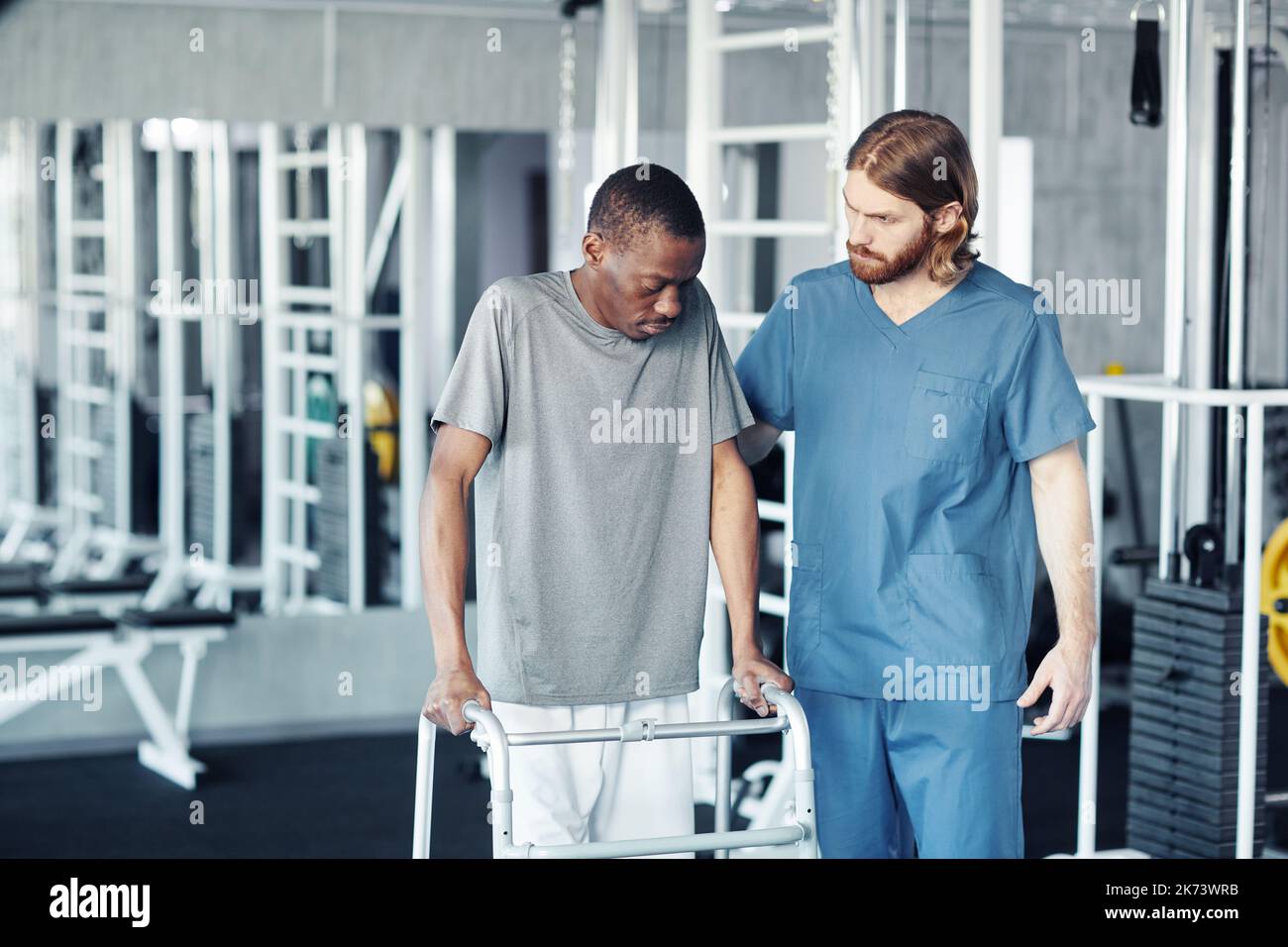 Young male doctor helping patient to move with walker during their exercises in gym Stock Photo