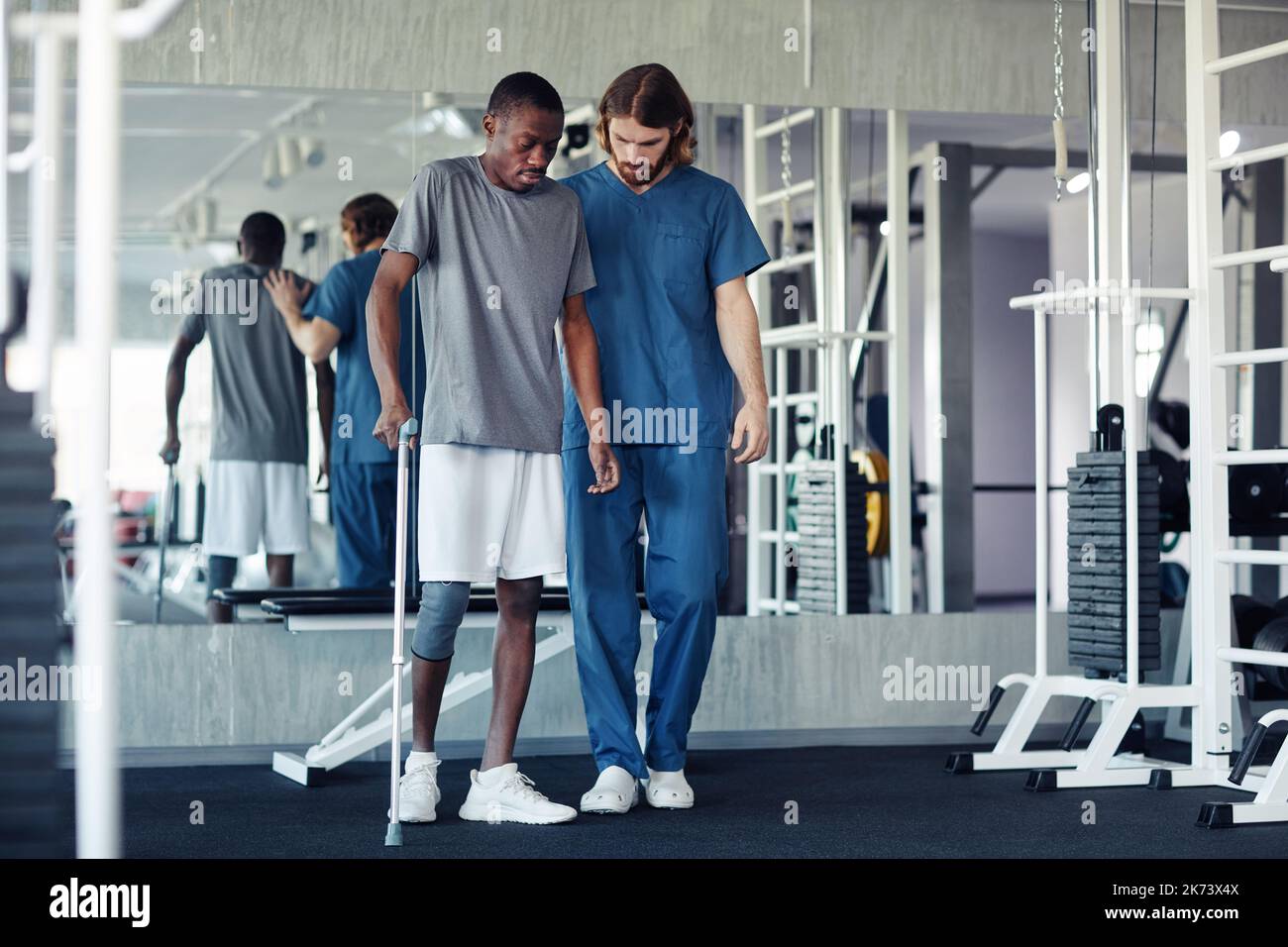 African patient trying to walk with his pain leg using walking stick with the help of doctor in gym Stock Photo