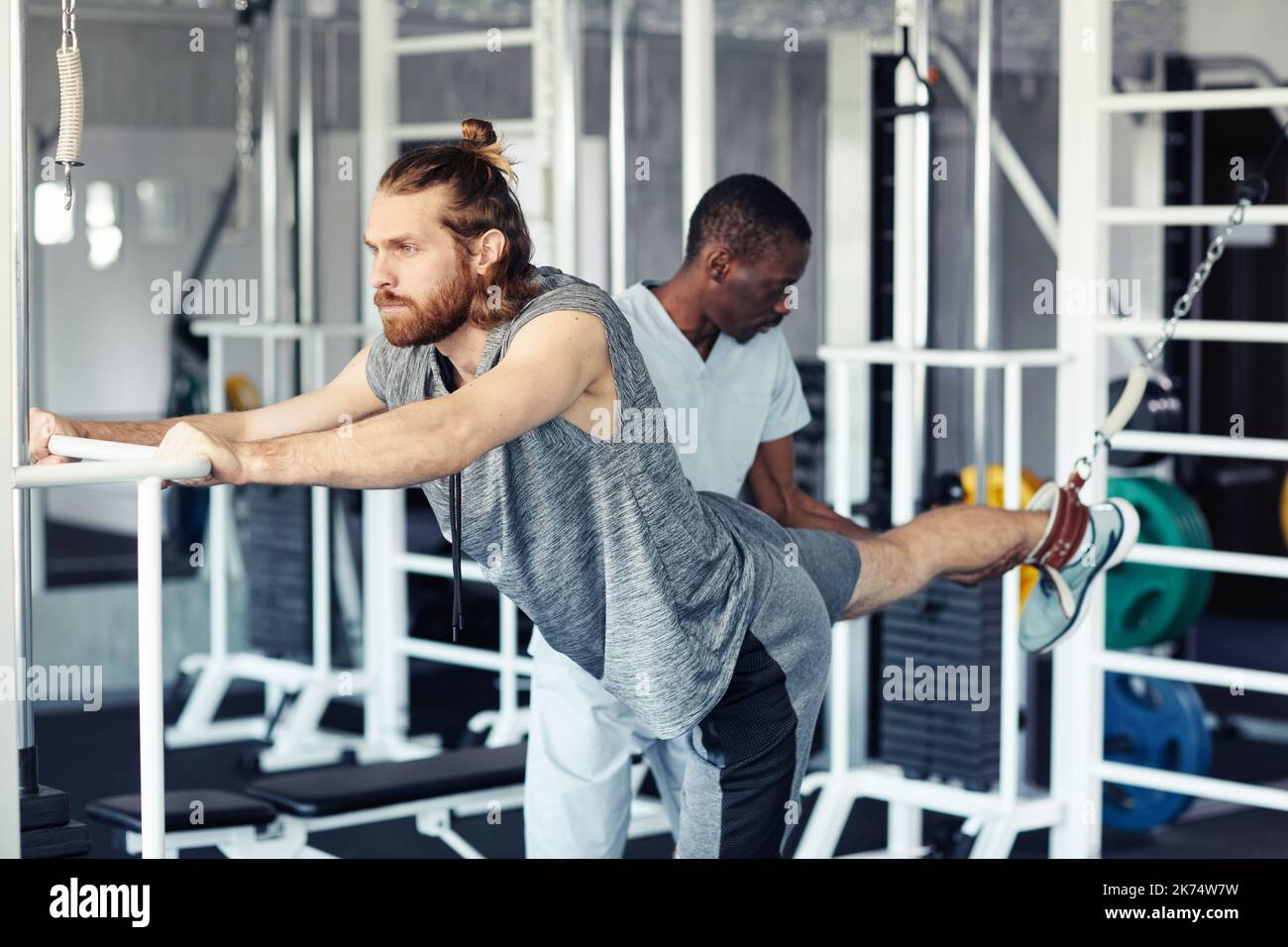 Young patient stretching his injury leg on sport equipment in gym with therapist helping him Stock Photo