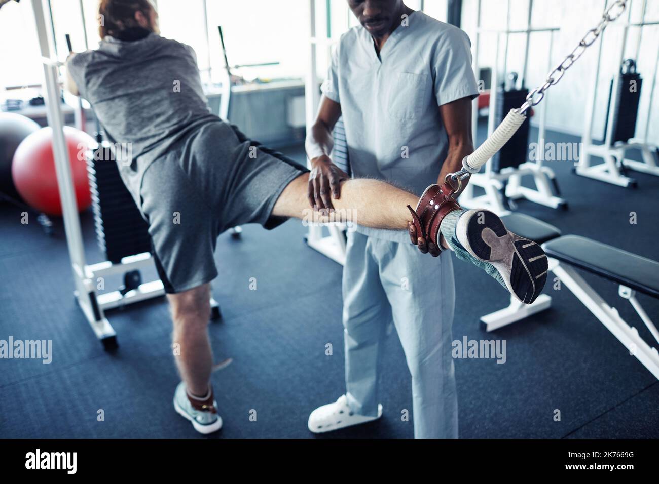 Rear view of young patient exercising on sport equipment to stretch his injury leg while holding rehabilitation with therapist in gym Stock Photo