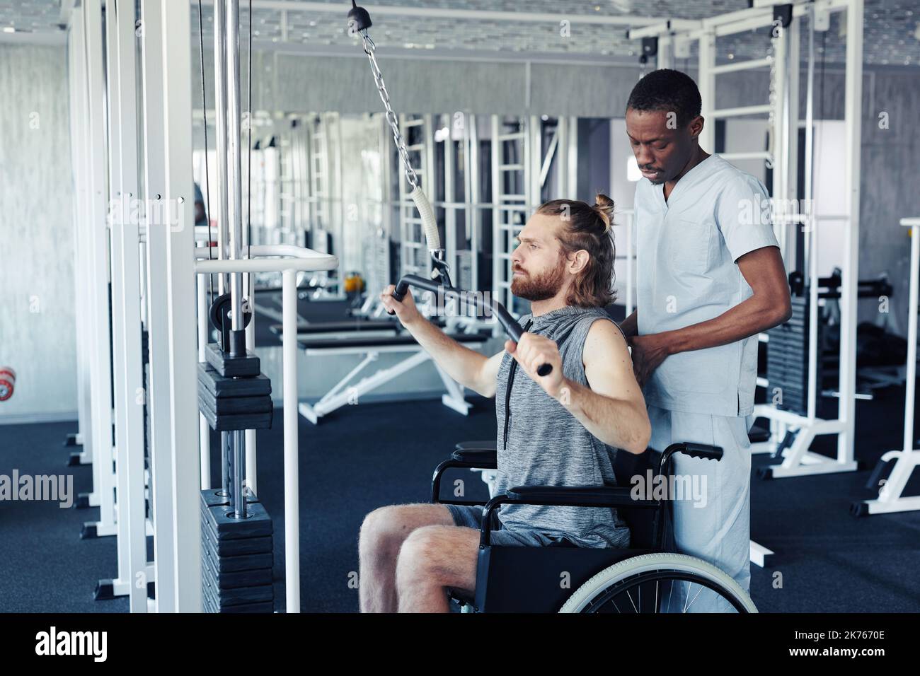 Patient with disability exercising on training equipment with therapist standing behind him and controlling his training in gym Stock Photo