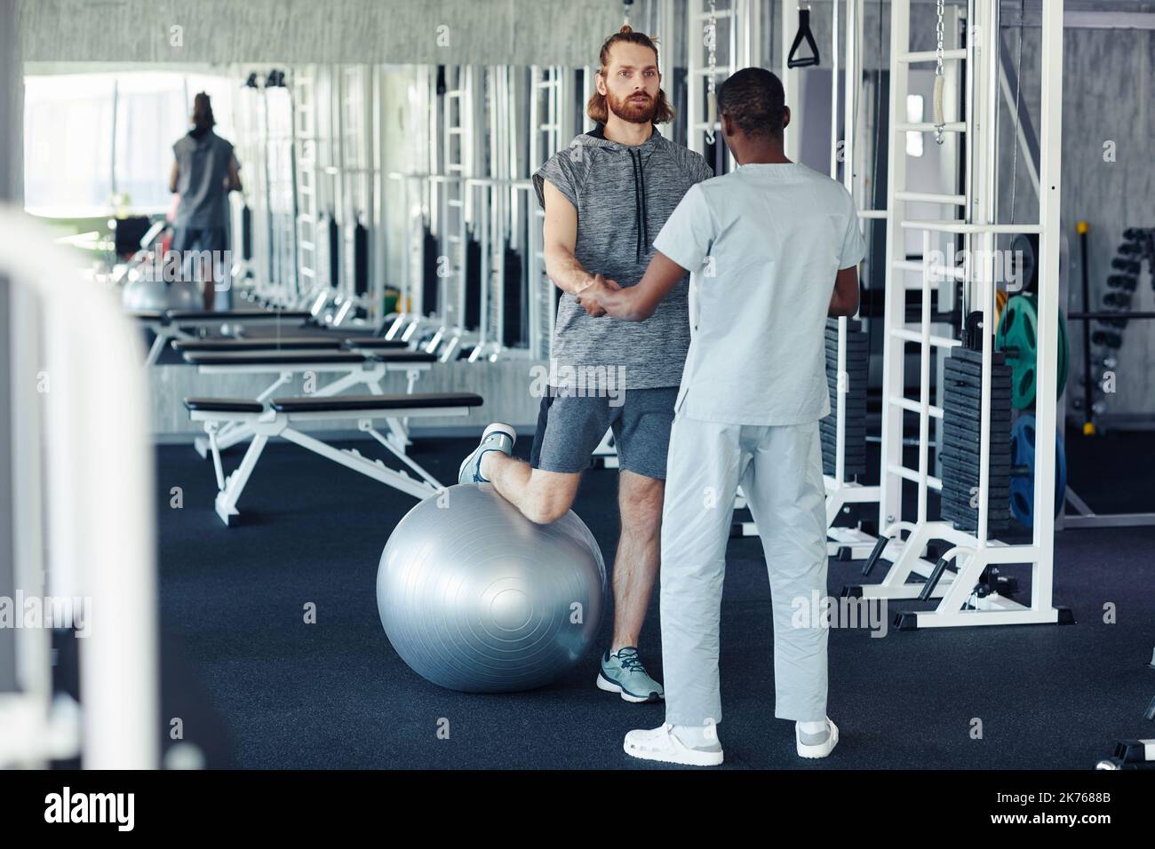 Patient doing exercises on fitness ball for recovering of his leg together with doctor in gym Stock Photo