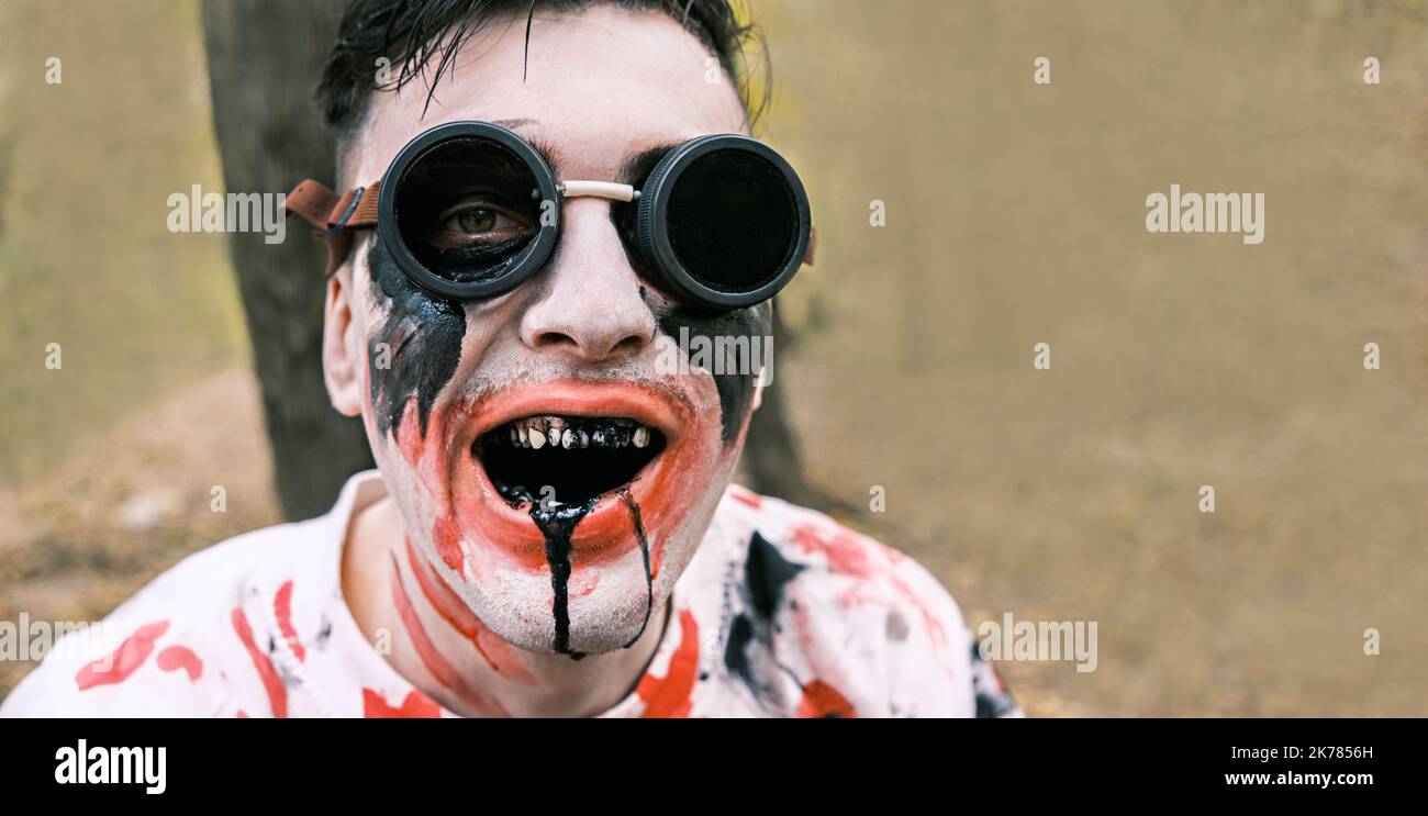 Portrait is close-up of face of zombie with traces of blood and black fluid flowing from the mouth. Man with makeup with aviation glasses with broken Stock Photo