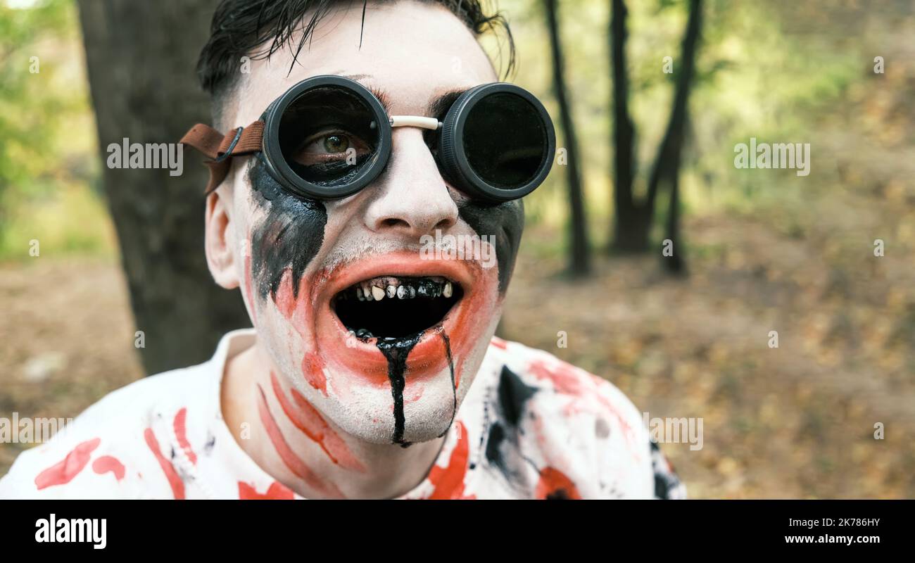 Portrait is close-up of face of zombie with traces of blood and black fluid flowing from the mouth. Man with makeup with aviation glasses with broken Stock Photo