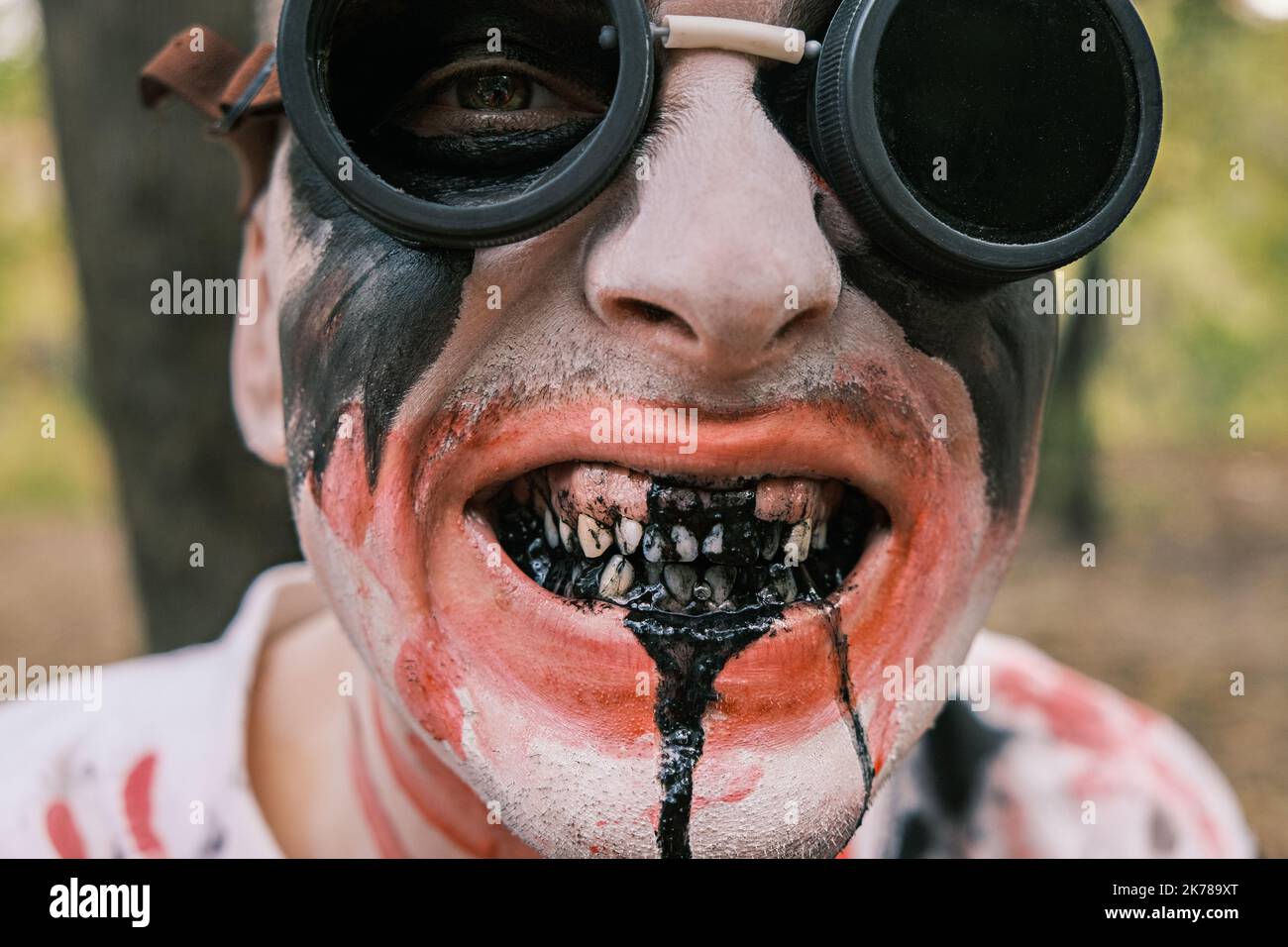 Close-up portrait of face of zombie with traces of blood and black fluid flowing from the mouth. Man with makeup with aviation glasses with broken Stock Photo