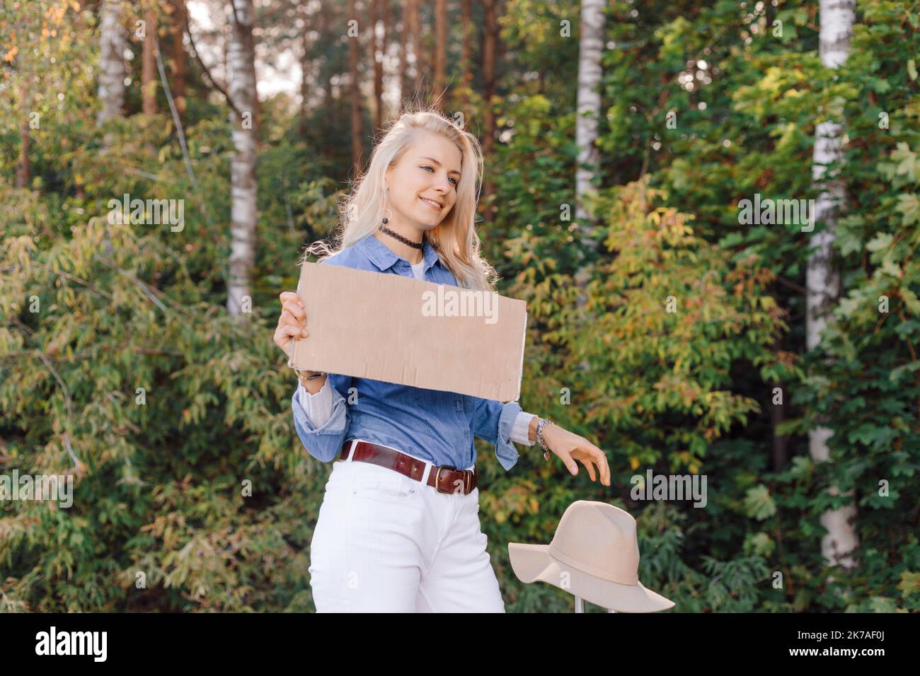 Pretty woman wait passing car in forest standing with suitcase and cardboard poster on roadside of highway. Blonde lady escape from city to go Stock Photo