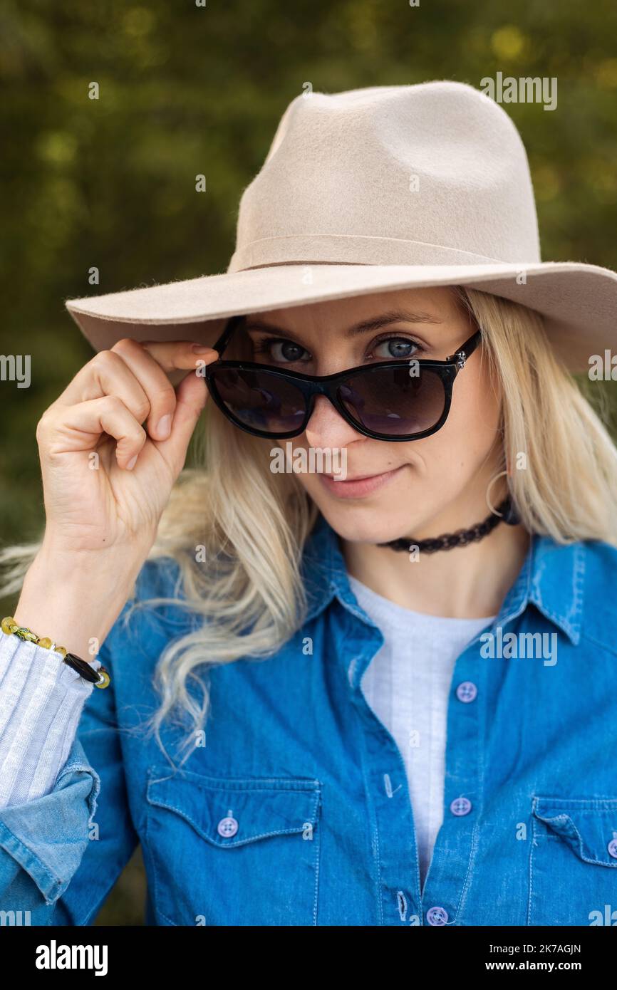 Portrait closeup of beautiful blonde woman in sunglasses and elegant hat on forest background. Stylish traveller in seasonal denim outfit. Autumn Stock Photo
