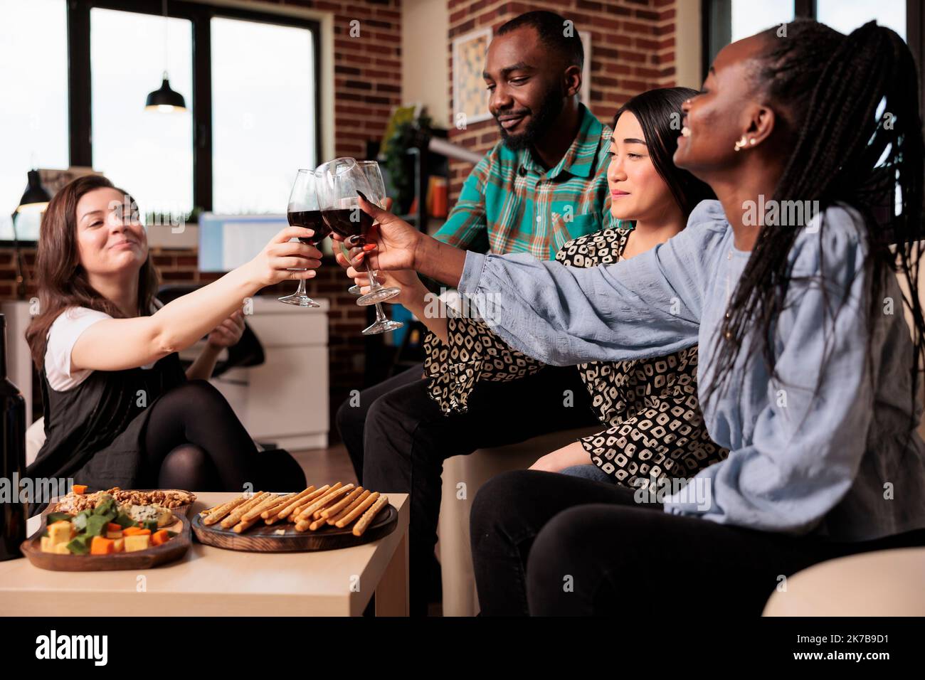 Different ethnicities friends group, toasting, drinking wine, eating snacks, having fun at young adult birthday living room apartment party. Multicultural people enjoying friendship. Stock Photo