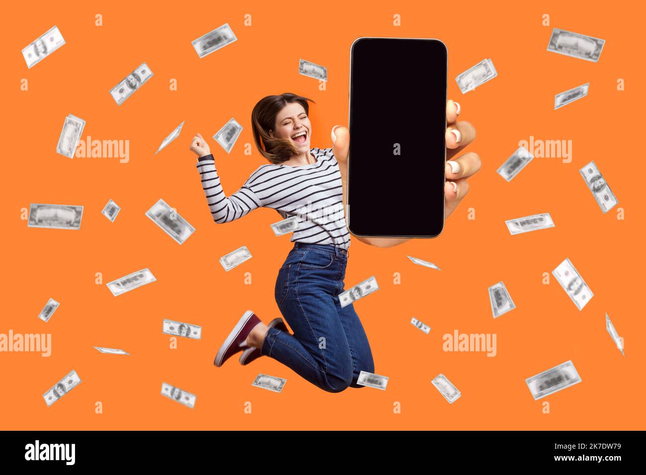 Portrait of extremely excited woman wearing striped shirt jumping in money rain showing phone display with advertisement area, betting and winning. Indoor studio shot isolated on orange background. Stock Photo