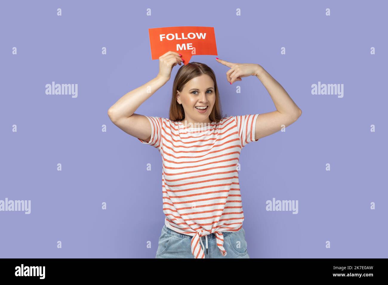 Portrait of delighted smiling blond woman holding red card with follow me inscription above head, pointing at poster, asking to subscribe. Indoor studio shot isolated on purple background. Stock Photo