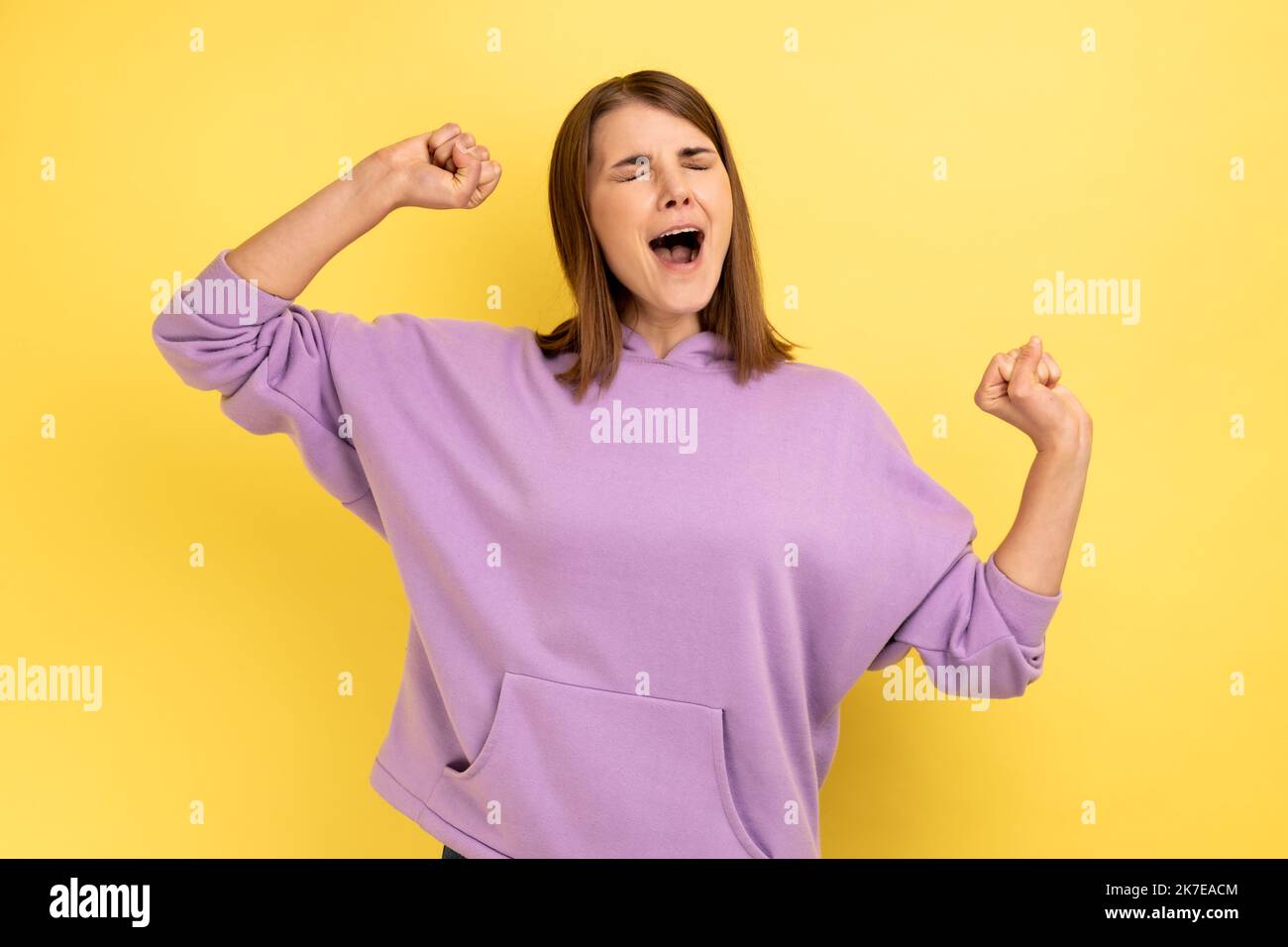 Portrait of young adult sleepless woman yawning and raising hands up, feeling fatigued, standing with close eyes, wearing purple hoodie. Indoor studio shot isolated on yellow background. Stock Photo