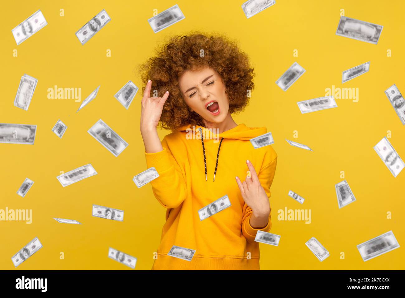 Portrait of woman with Afro hairstyle wearing casual style hoodie showing rock and roll, rejoicing money rain falling from up, winning lottery. Indoor studio shot isolated on yellow background. Stock Photo
