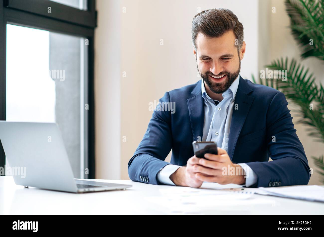 Online messaging. Positive busy caucasian business man in a suit, company employee, top manager, sitting in the office, using his smart phone, texting online with colleague or client, smiling Stock Photo