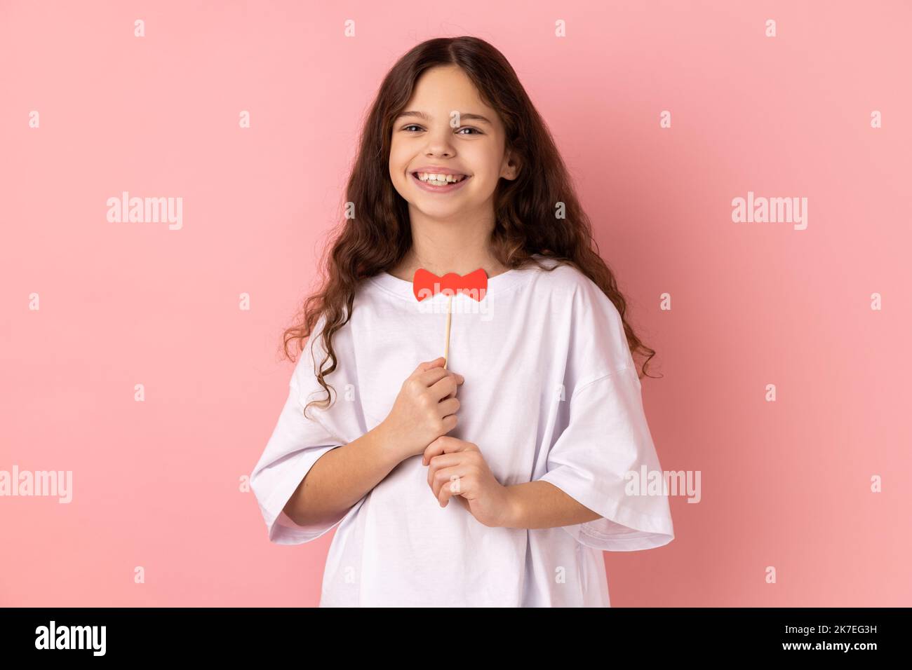 Portrait of satisfied little girl wearing white T-shirt holding funny paper bow tie, having fun playing game, wearing masquerade accessory. Indoor studio shot isolated on pink background. Stock Photo