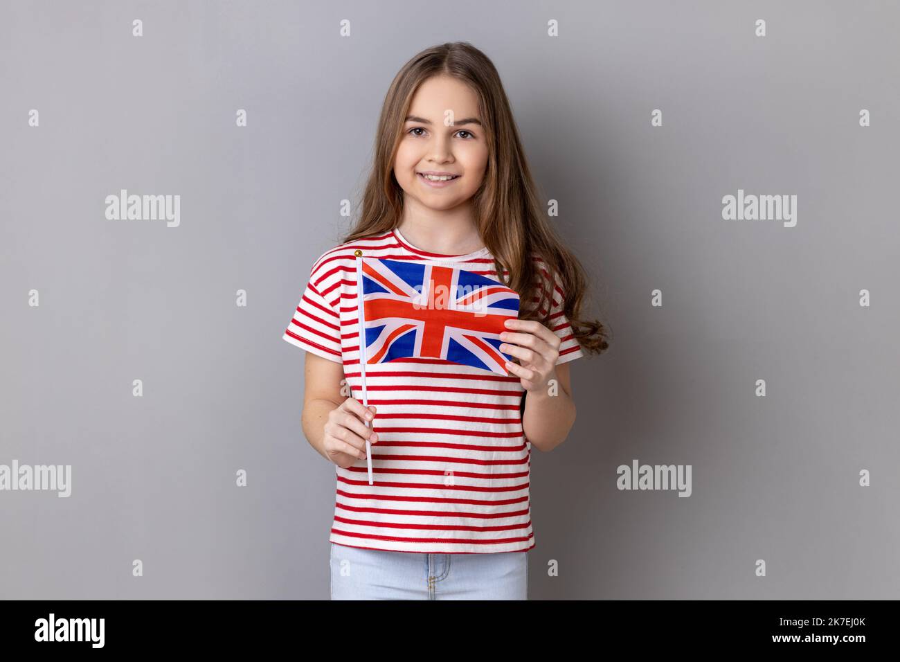 British flag. Portrait of charming smiling little girl wearing striped T-shirt holding flag of United Kingdom looking at camera with positive expression. Indoor studio shot isolated on gray background Stock Photo