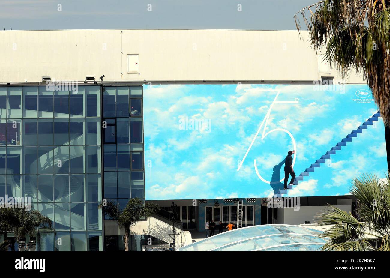 ©PHOTOPQR/NICE MATIN/Patrice Lapoirie ; Cannes ; 16/05/2022 ; Le rituel traditionnel vient d’avoir lieu ce dimanche matin. L’affiche du 75e Festival de Cannes, dont l’image est extraite du film Truman Show, a été hissée et fixée sur le frontispice du Palais. C’est parti pour la Quinzaine du 7è art ! Un homme gravit des marches célestes, qui le mènent vers les nuages, et touche du doigt le '5' de la 75è édition du Festival du film. Allégorie d’un cinéma qui reste synonyme d’évasion et de liberté créatrice, dans un monde où ces valeurs sont parfois menacées? Quoi qu’il en soit, l’affiche du Fes Stock Photo