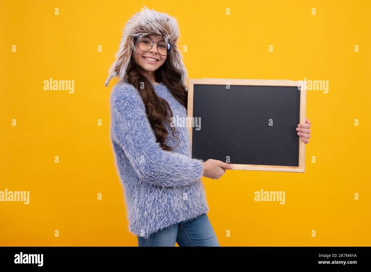 Teenage girl child in warm autumn hat and sweater holding blackboard, isolated on a yellow background. Autumn school sale, copy space, mockup. Happy Stock Photo