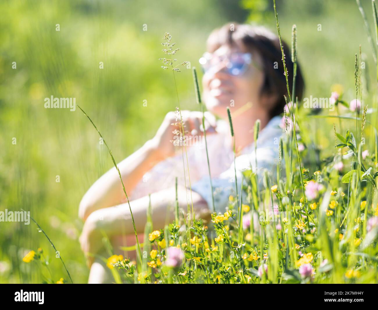 Blurred background with woman in sunglasses. Woman enjoys sunlight and flower fragrance on grass field. Summer vibes. Relax outdoors. Self-soothing. Stock Photo