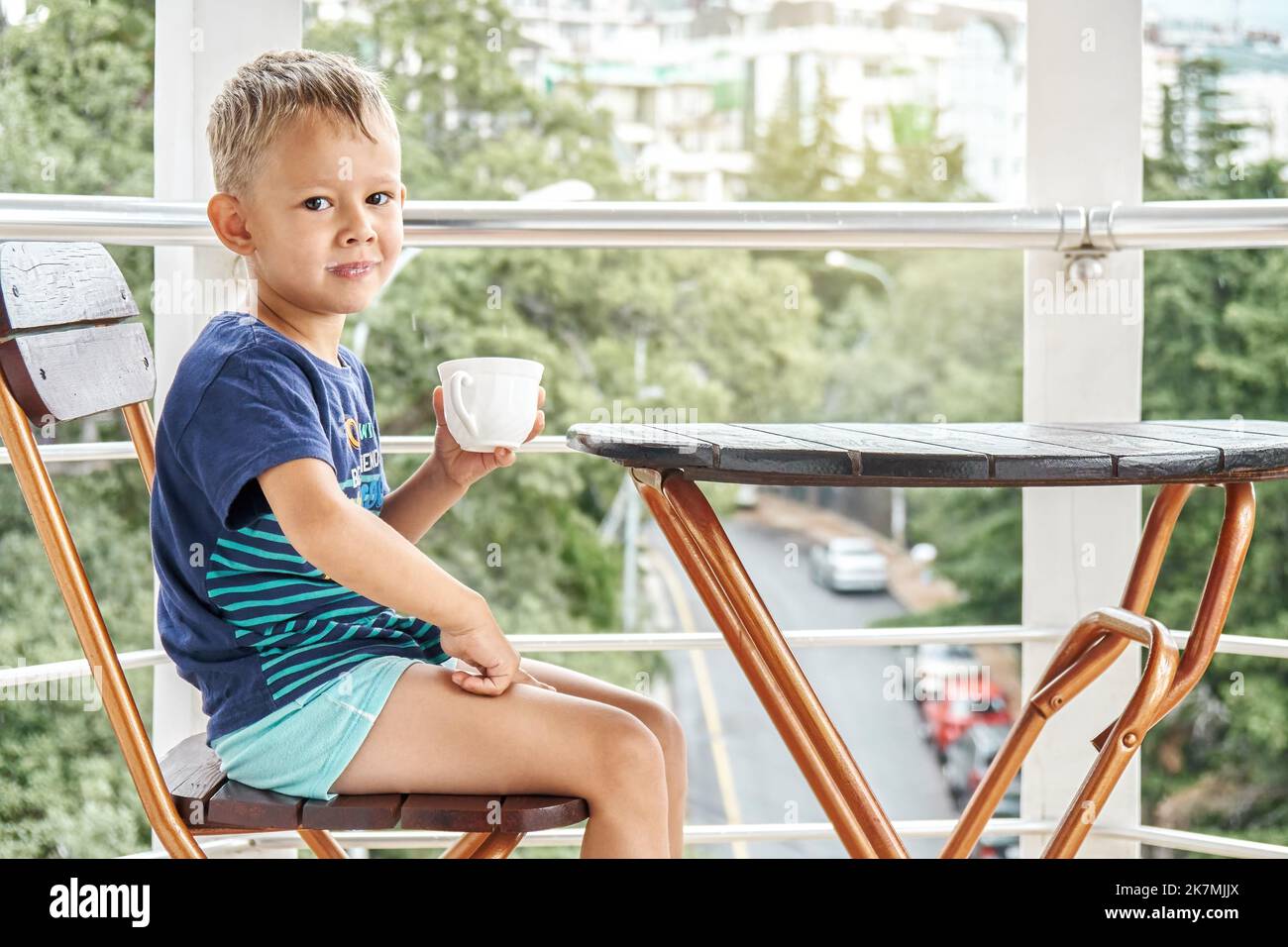Preschooler boy drinks milk from mug sitting on chair on hotel balcony