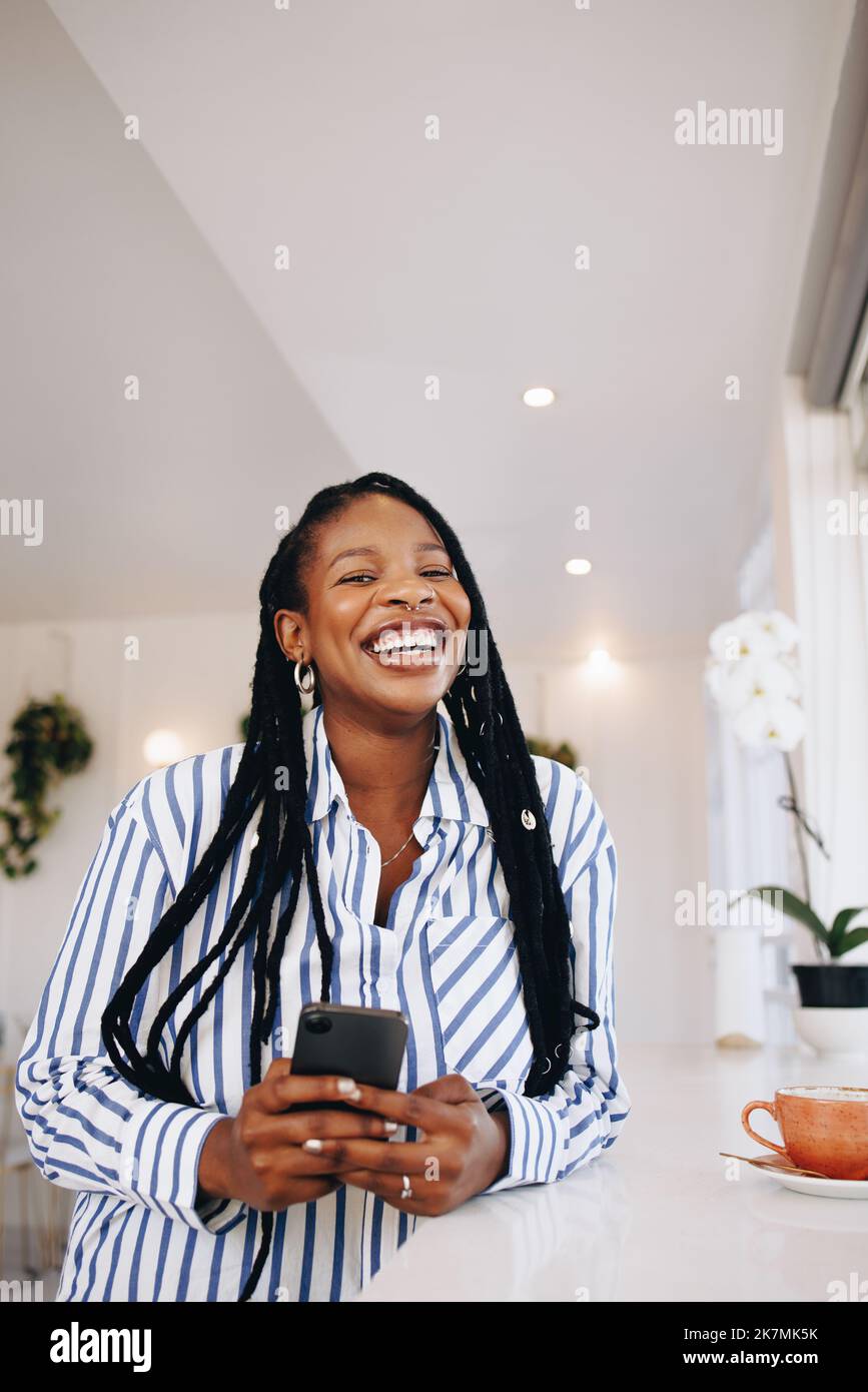 Portrait of a young businesswoman laughing happily while holding a smartphone in a coffee shop. Cheerful young black businesswoman enjoying her coffee Stock Photo