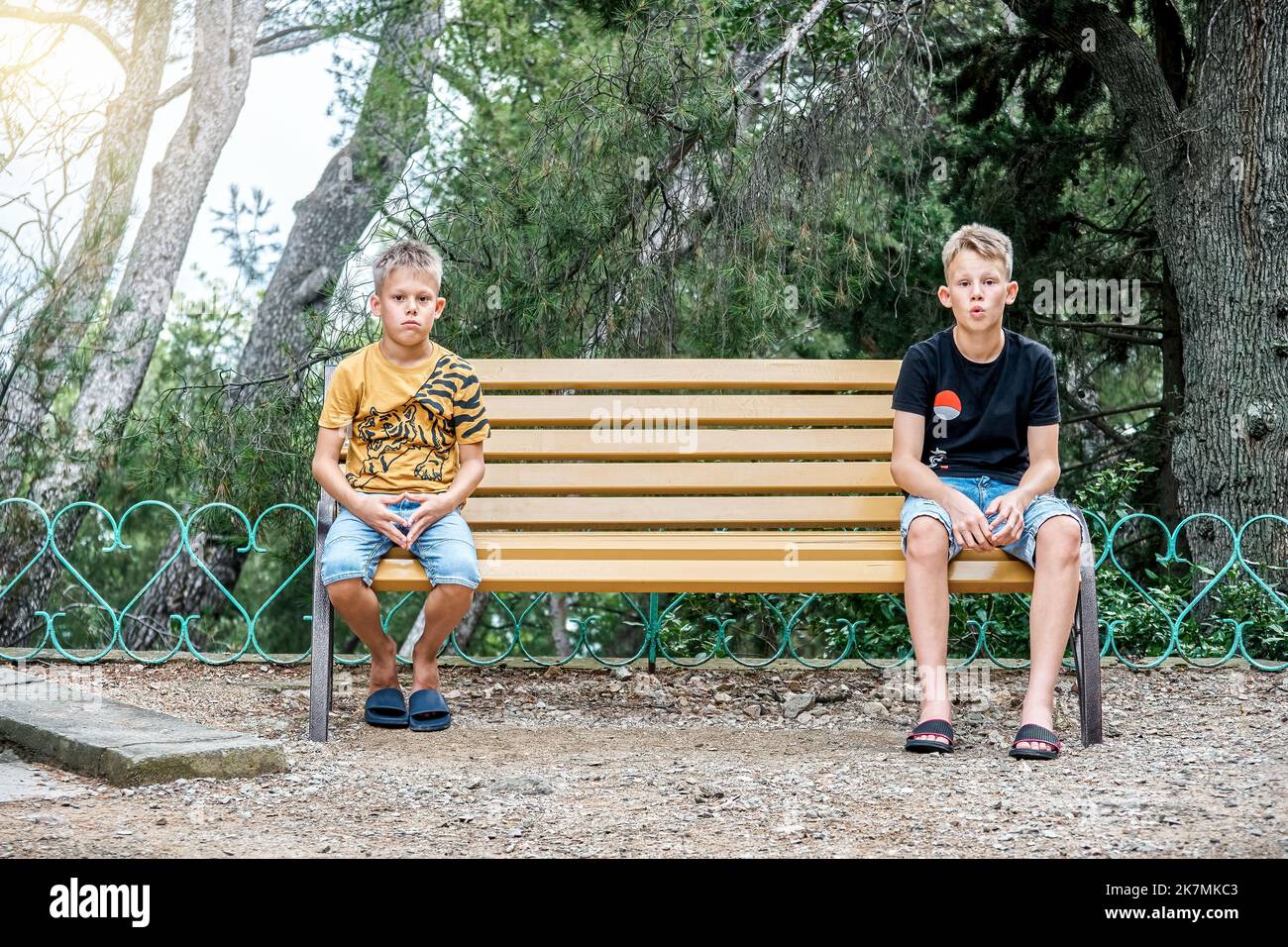 Siblings sitting on wooden bench look with upset and disappointed expressions. Brothers sit on different sides of bench in silence after argument Stock Photo